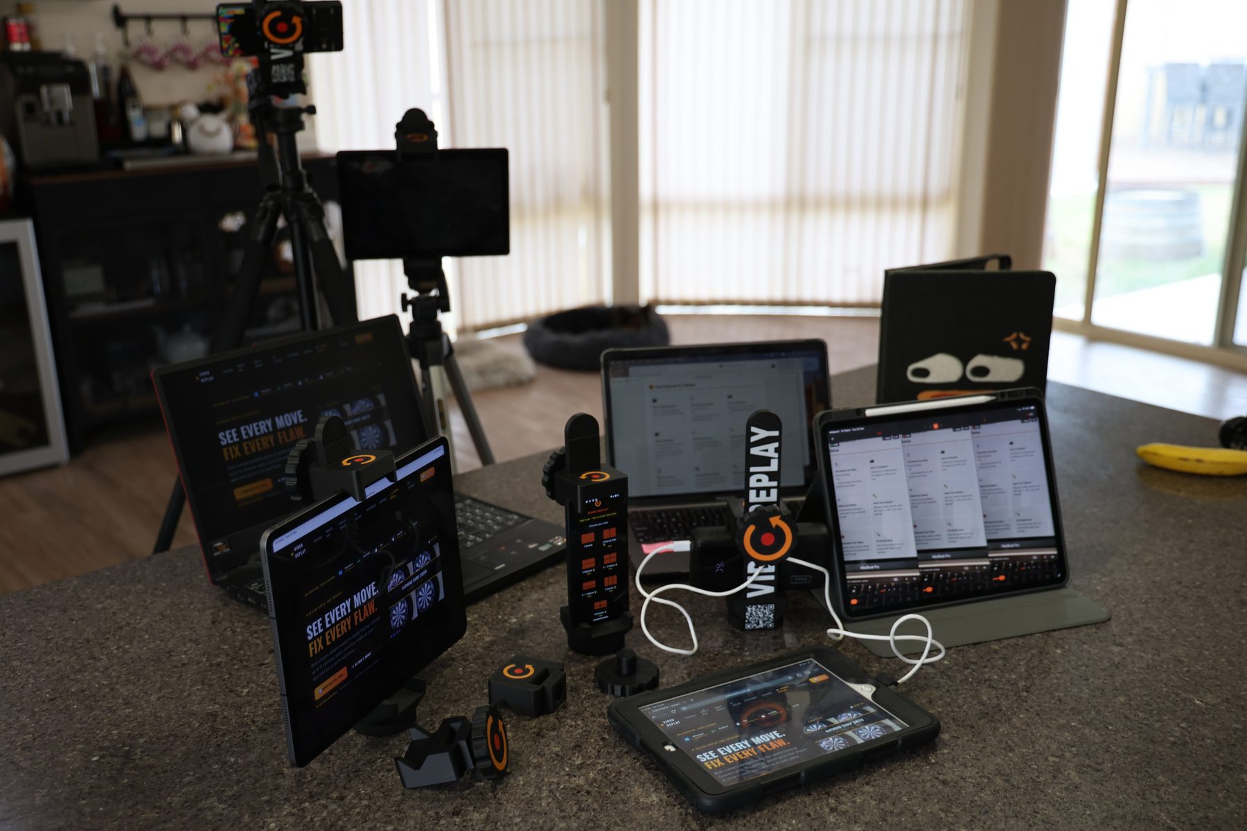 A lineup of Video Replay tripod holders, phone and tablet editions, mounted on tripods and holding phones, tablets and a MacBook running the Video Replay app